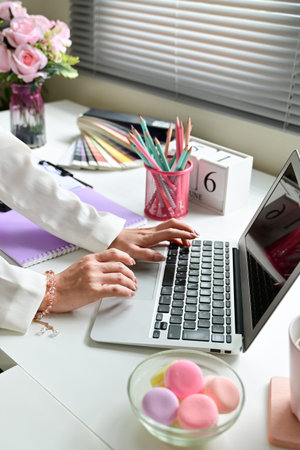 A woman is typing on a laptop in front of a white desk. The desk is cluttered with various items...