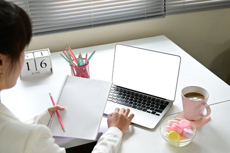 A woman is sitting at a desk with a laptop and a notebook. She is writing in the notebook and typing on the laptop. The scene suggests that she is working or studyingの写真素材