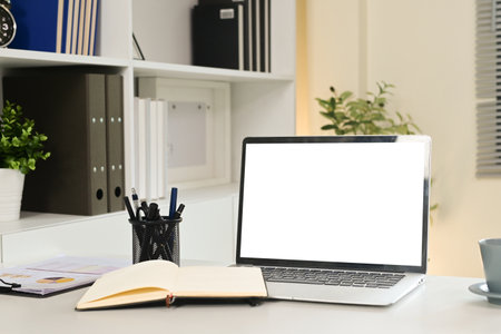 A laptop sits on a desk with a notebook and a cup of coffee. The laptop is open to a blank screen.の写真素材