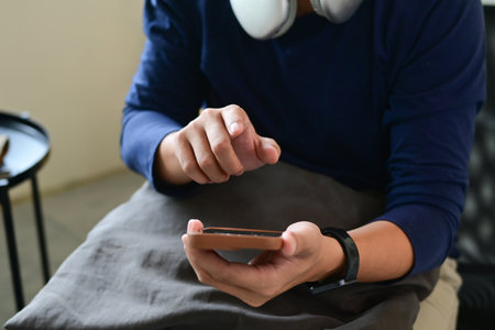 A man is sitting on a couch, holding a cell phone in his hands and looking at it intently. He is focused on the screen, possibly checking his messages or browsing the internet.の写真素材