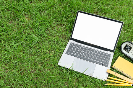 A laptop is on a grassy field with a clock and a notebook. The laptop is open and the grass is greenの写真素材