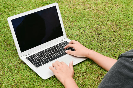 A person is typing on a laptop in a grassy field. The laptop is open and the screen is blackの写真素材