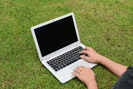 A person is typing on a laptop in a grassy field. The laptop is open and the screen is black.の写真素材