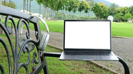 A laptop is sitting on a bench in a park. The laptop is open and the screen is blank. The bench is located in a grassy area with trees in the background.の写真素材