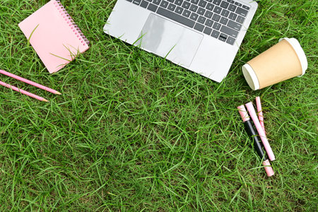 A laptop, a cup of coffee, and a few pens are on a grassy field. The scene suggests a relaxed and...