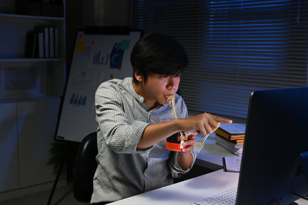 A man is eating a piece of Instant noodles while pointing at a computer monitor..の写真素材