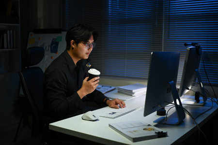 A man is sitting at a desk with a cup of coffee in front of him. He is wearing glasses and is typing on a computer. Concept of focus and productivityの写真素材