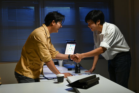 Two men shaking hands in a dimly lit room. One of them is wearing glasses. Scene is professional and friendlyの写真素材