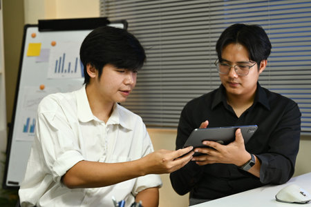 Two men are sitting at a desk with a computer monitor in front of them. One of the men is holding a tablet. They are discussing something on the tablet.の写真素材