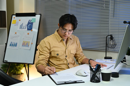A man in a shirt is sitting at a desk with a white board behind him. He is writing on a piece of paper with a pen.の写真素材