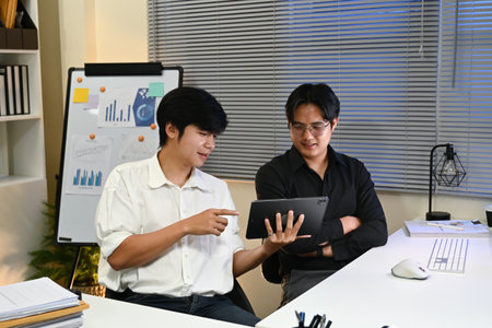 Two men are sitting at a desk with a white board behind them. One of the men is holding a tablet and pointing at something on the board. Scene is focused and professional.の写真素材