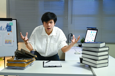 A man is sitting at a desk with a stack of books and a computer. He is angry and frustrated, possibly due to a work-related issueの写真素材