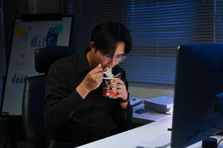 A man is eating noodles in front of a computer monitor. He is wearing glasses and a black shirtの写真素材