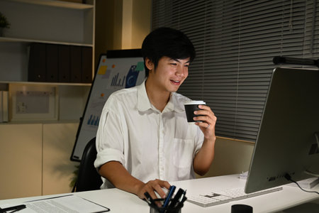 A man is sitting at a desk with a computer monitor and a cup of coffee. He is smiling and he is enjoying his coffee while working.の写真素材