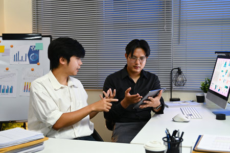 Two men are sitting at a desk with a computer monitor in front of them. One of the men is holding a tablet. They are discussing something on the tabletの写真素材