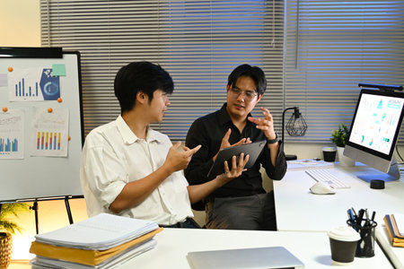Two men are sitting at a desk with a white board behind them. One of the men is holding a tablet and pointing at something on the board. Scene is focused and professionalの写真素材