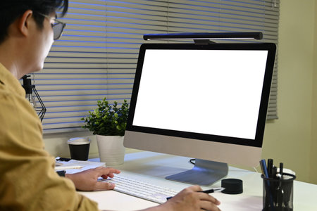 A person is typing on a computer monitor with a white background. The person is wearing glasses and is sitting at a desk with a potted plant in front of them..の写真素材