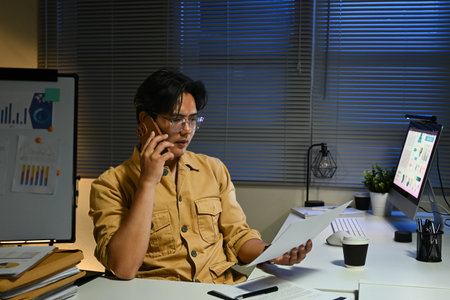A man in a yellow shirt is talking on his cell phone while looking at a piece of paper. He is sitting at a desk with a computer monitor and a keyboard. The room is dimly lit.の写真素材