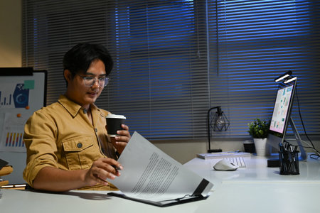 A man is sitting at a desk with a cup of coffee and a piece of paper in front of him. He is reading the paper and sipping his coffee.の写真素材