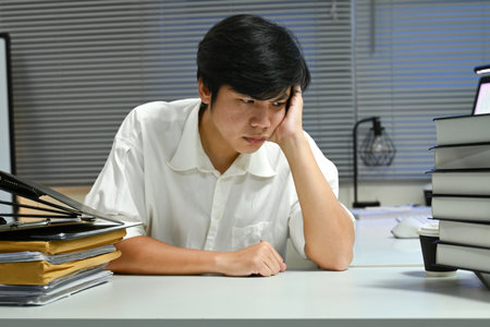 A man is sitting at a desk with a stack of books and papers in front of him. He is looking down and he is in a state of deep thoughtの写真素材