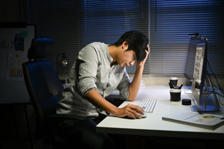 A man is sitting at a desk with a computer monitor and a keyboard. He is looking at the monitor and he is in a state of distressの写真素材
