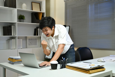 A man is sitting at a desk with a laptop and a cup of coffee. He is smiling and he is happyの写真素材