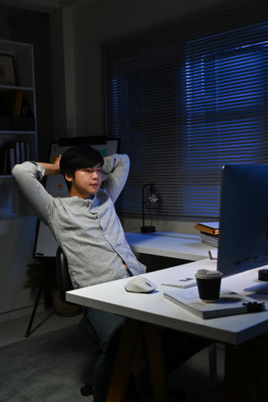 A young man is sitting at a desk with a computer monitor in front of him. He is wearing a white shirt and he is in a relaxed state.の写真素材