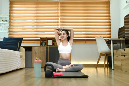 A woman is doing a yoga pose in her living room. She is smiling and she is enjoying herself. There are several items in the room, including a bottle, a cup, a potted plant, and a couch.の写真素材