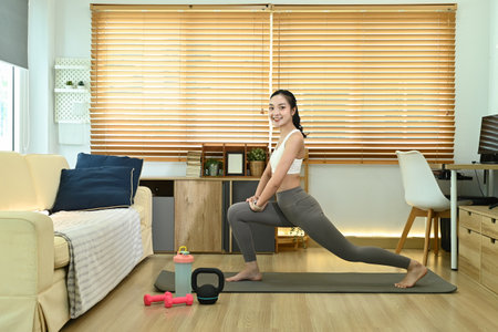 A woman is doing a yoga pose in a living room. She is wearing a white tank top and grey pants. There is a kettlebell and a pair of dumbbells on the floor next to herの写真素材