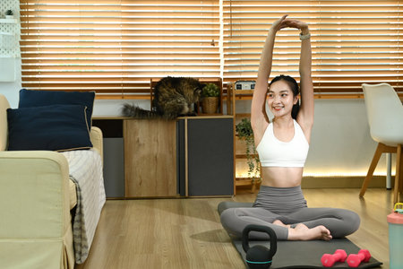 A woman is doing yoga in her living room. She is wearing a white tank top and gray pants. A cat is sitting on a couch in the background.の写真素材