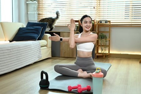 A woman is doing yoga in her living room. She is wearing a white tank top and gray pants. A cat is sitting on a couch in the background.の写真素材