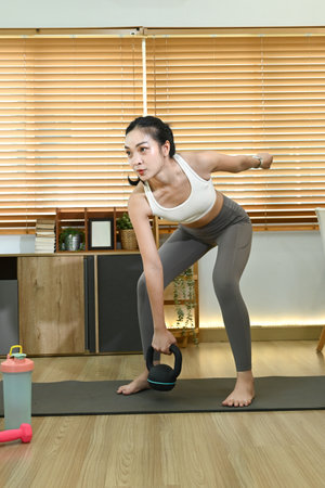 A woman is doing a workout in her living room. She is holding a kettlebell and is wearing a white tank top. The room has a couch and a few potted plants.の写真素材