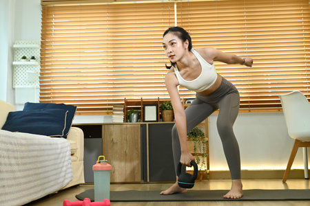 A woman is doing a workout in her living room. She is holding a kettlebell and is wearing a white tank top. The room has a couch and a few potted plants.の写真素材