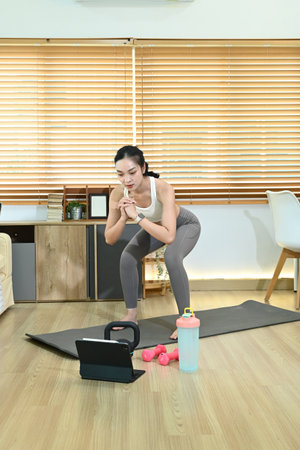 A woman is doing a workout in her living room. She is using a tablet to follow a workout video. There are two dumbbells and a water bottle nearby. The room has a wooden floorの写真素材