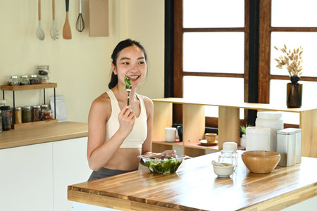 A woman is eating a salad in a kitchen. The kitchen is well-equipped with various utensils and bowls.の写真素材