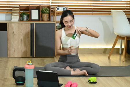 A woman is sitting on a mat with a tablet in front of her. She is drinking a green smoothie and has a bottle of protein powder next to her. Concept of health and wellnessの写真素材