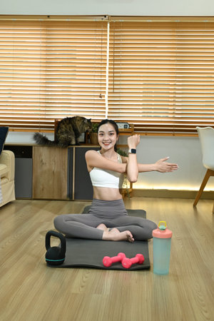 A woman is doing yoga in her living room. She is wearing a white tank top and gray pants. A cat is sitting on a couch in the background.の写真素材