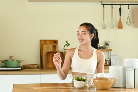 A woman is smiling and holding a fork with a salad in front of her. The table is set with bowls, cups, and spoons, and there are several bottles and jars on the table.の写真素材