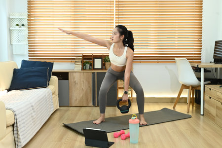 A woman is doing a workout in her living room. She is holding a kettlebell and is wearing a white tank top. The room has a couch and a few potted plants.の写真素材