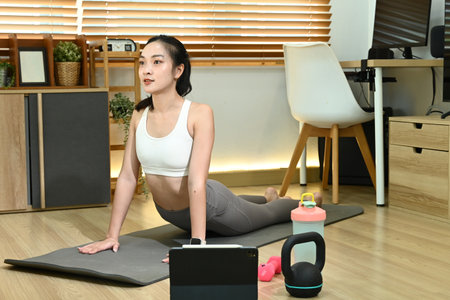 A woman is doing yoga in her living room. She is using a laptop and a pair of dumbbells.の写真素材