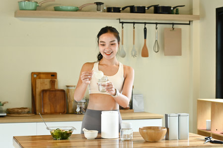A woman is in a kitchen, smiling and preparing a drink. The kitchen is well-equipped with various utensils and appliances, including a blender, a bowl, a spoon, and a cup.の写真素材