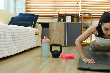 A woman is doing a push up on a mat in a living room. There is a kettlebell and a water bottle nearbyの写真素材