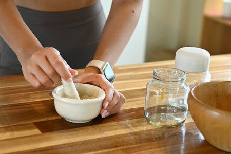 A woman is preparing a mixture in a bowl on a wooden table. The table is cluttered with jars, a bowl, and a bottle. The woman is focused on her task, and the scene conveys a sense of concentration.の写真素材