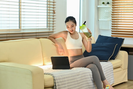A woman is sitting on a couch with a laptop and a green bottle in her hand. She is smiling and she is enjoying her timeの写真素材