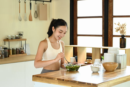 A woman is eating a salad in a kitchen. The kitchen is well-equipped with various utensils and bowlsの写真素材