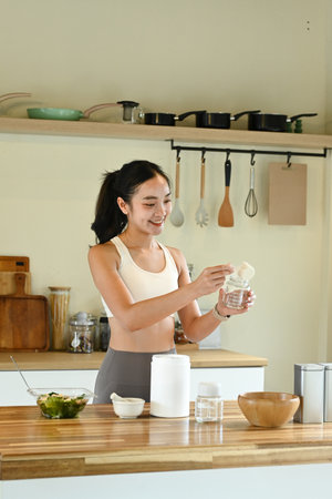A woman is in a kitchen, smiling and preparing a drink. The kitchen is well-equipped with various utensils and appliances, including a blender, a bowl, a spoon, and a cupの写真素材