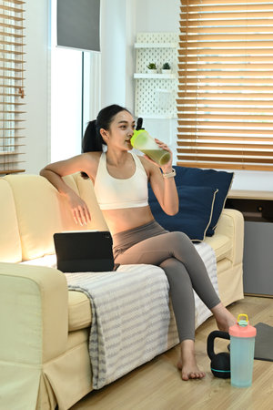 A woman is sitting on a couch and drinking a green smoothie. She is wearing a white tank top and grey pants. The room has wooden furniture and a laptop on the couch.の写真素材