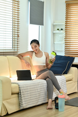 A woman is sitting on a couch with a laptop and a green bottle in her hand. She is smiling and she is enjoying her time.の写真素材