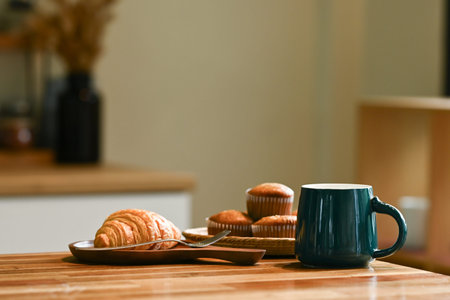 A plate of croissants and muffins sits on a wooden table next to a cup of coffee.