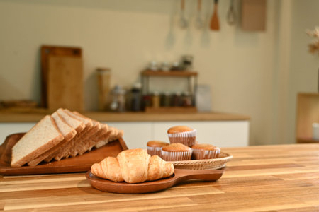 A plate of croissants and muffins sits on a wooden table. The table is surrounded by various kitchen items, including a knife, a spoon, and a bowl. The scene conveys a cozy and inviting atmosphereの写真素材